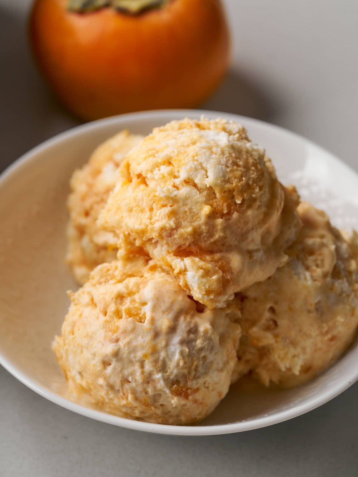 Four scoops of orange semifreddo in a small white bowl next to a persimmon.