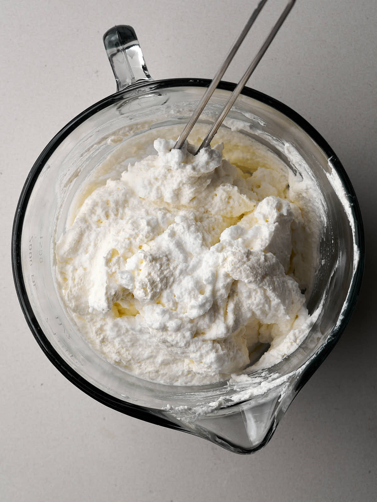 Whipped cream being mixed into meringue in a large glass bowl with a spout and handle.