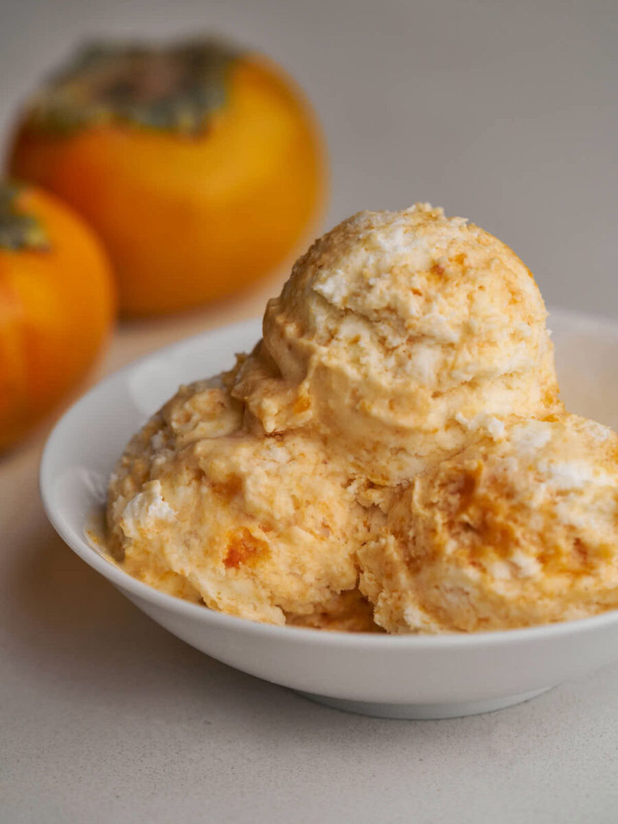 Three scoops of orange semifreddo in a small white bowl next to two persimmons.