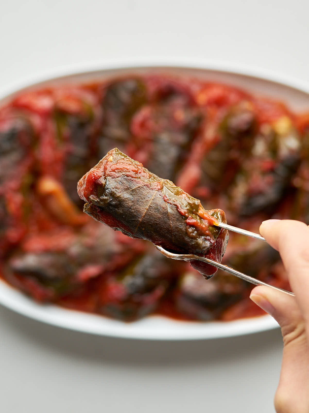 One baked dolma held in silver serving tongs above an oval plate of dolmas.