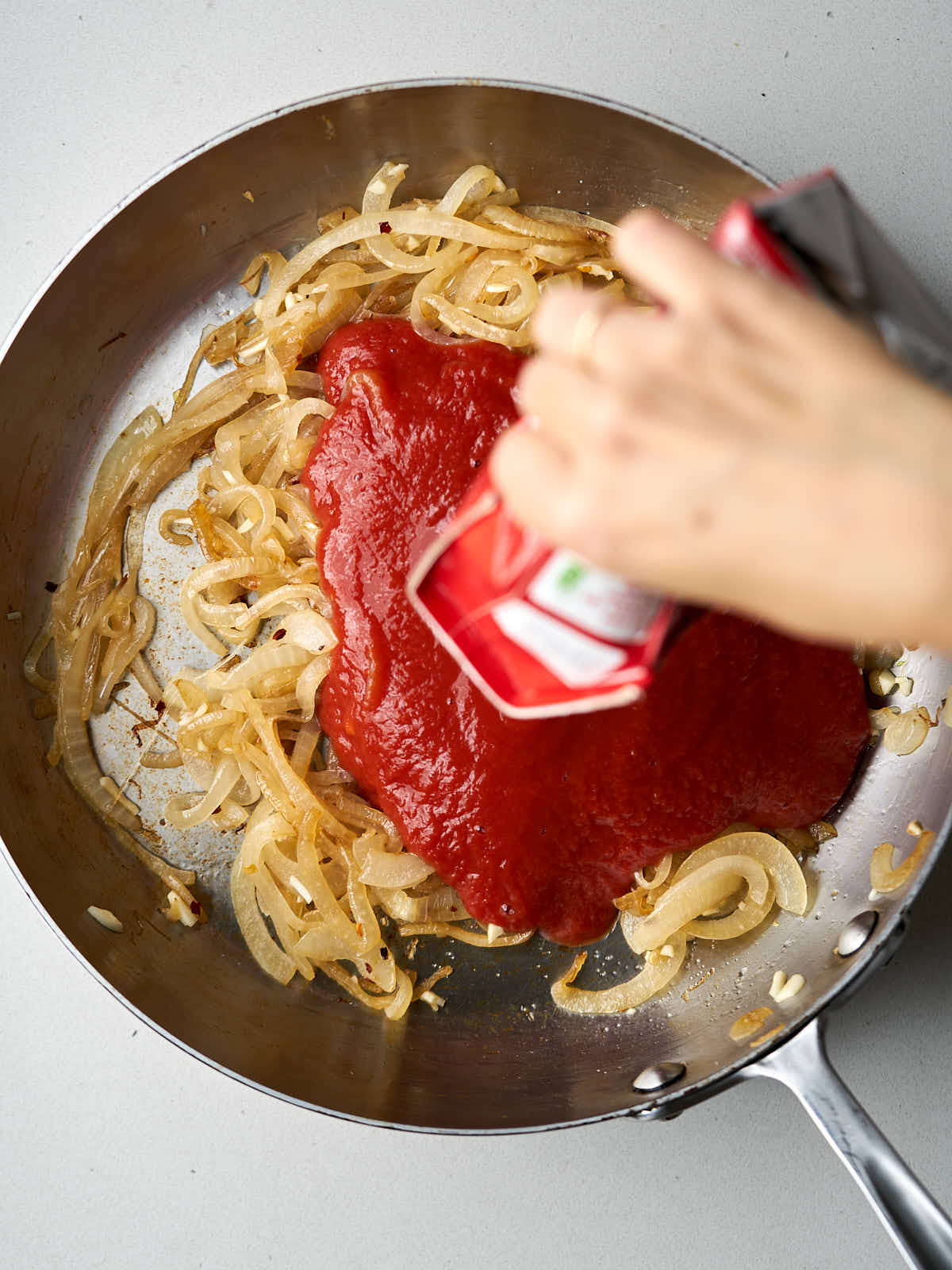 A hand pouring a can of tomato sauce over a pan with sauteed onions.