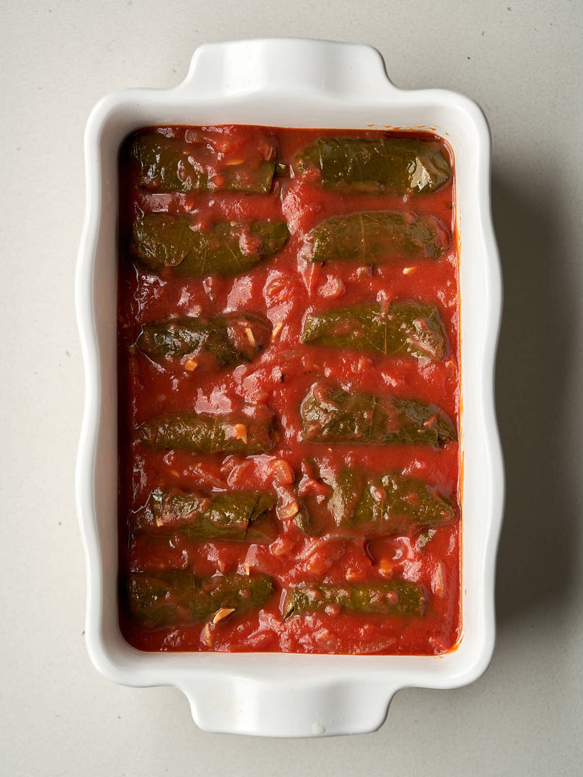 Wrapped, stuffed grape leaves submerged in tomato sauce in a ceramic dish.