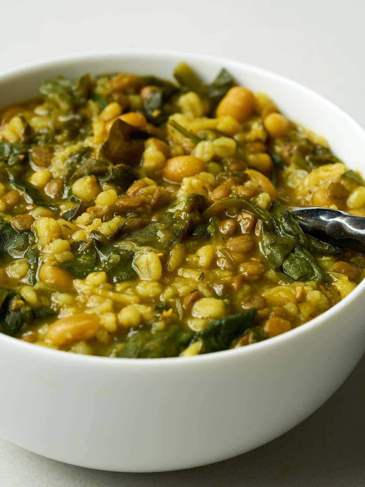 A bowl of mostly yellow and brown bean soup with cooked greens in it and a silver spoon coming out of it.