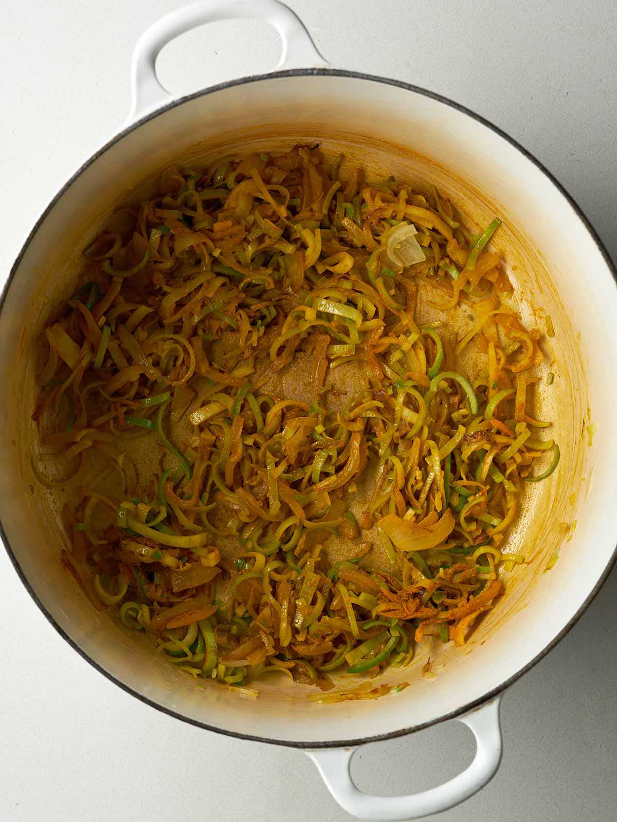 Sliced onions and leeks cooking in a white pot with yellow spices.