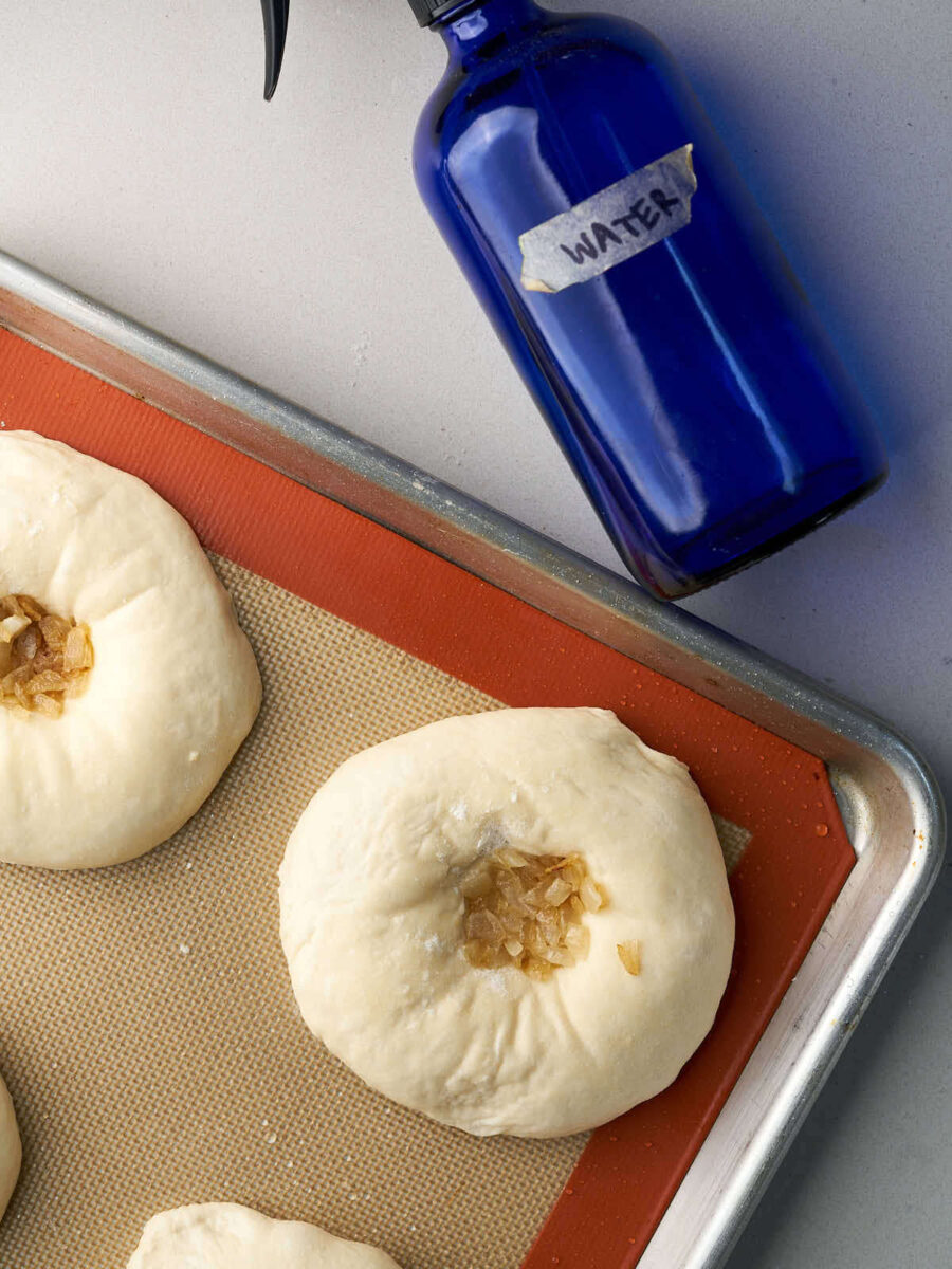 Bialys on a baking sheet next to a spray bottle with water.