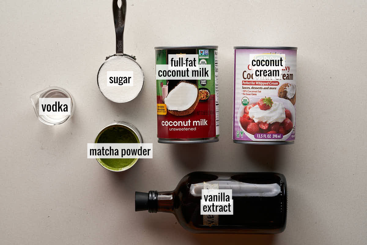 Two cans of coconut milk and cream next to matcha powder, an amber bottle, sugar, and a small beaker of vodka.