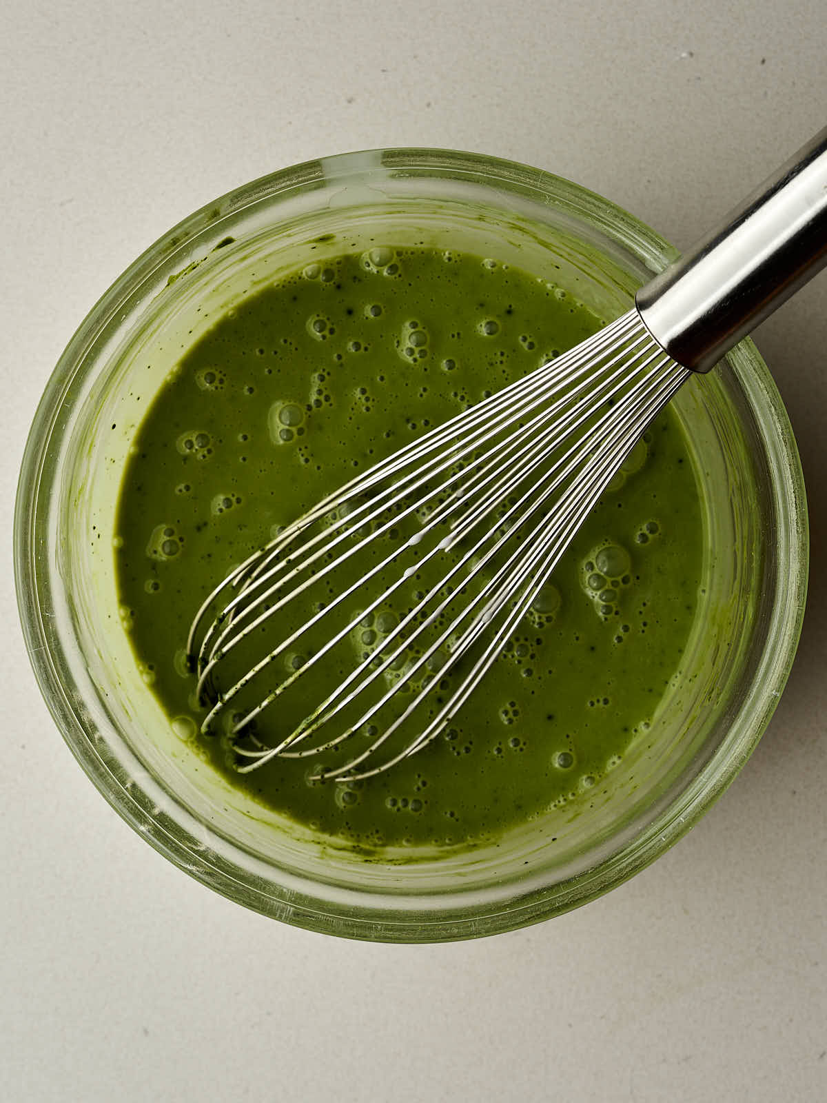 Thick green liquid with small bubbles in a glass bowl with a large metal whisk.