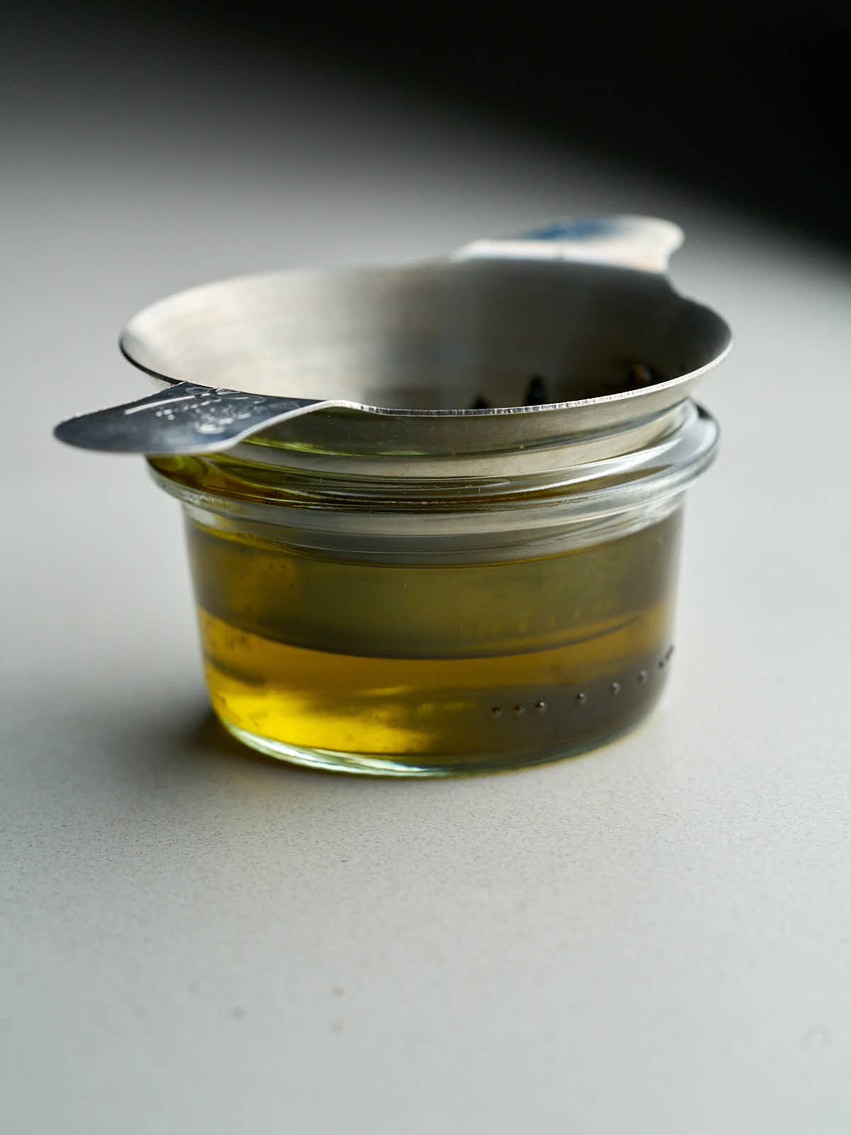 Small jar with olive oil topped with a metal strainer.