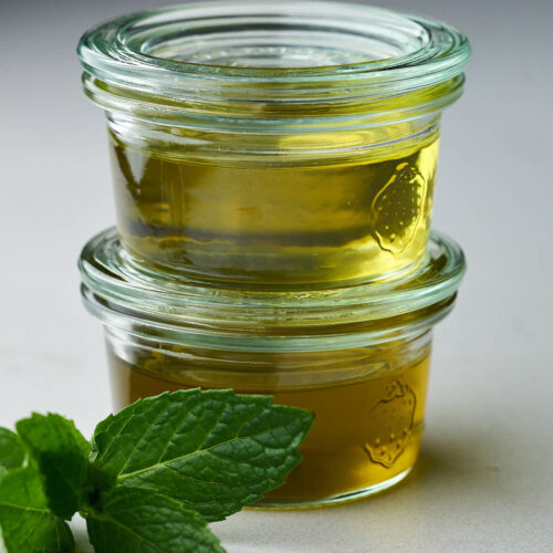 Two small jars of different colored oil stacked on top of each other next to a sprig of mint.