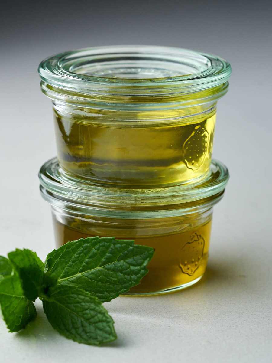 Two small jars of different colored oil stacked on top of each other next to a sprig of mint.