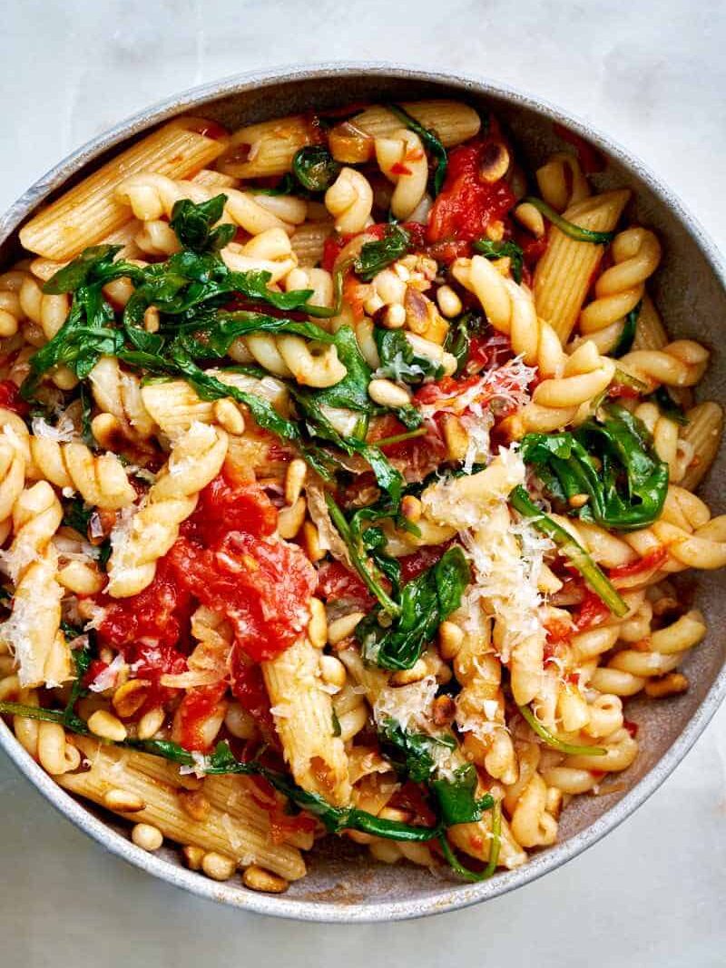 Close up of a bowl of pasta with arugula and tomato mixed in.