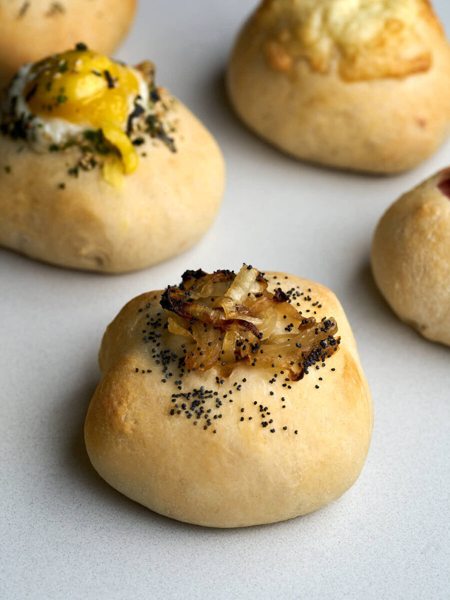 Bialy topped with caramelized onions and poppy seeds in front of bialys with other toppings.