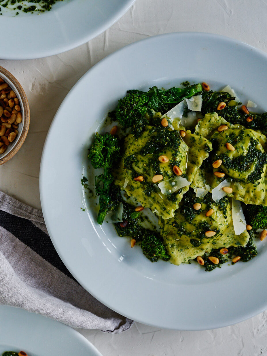 Three white plates with pesto-covered ravioli next to a small bowl od pine nuts.