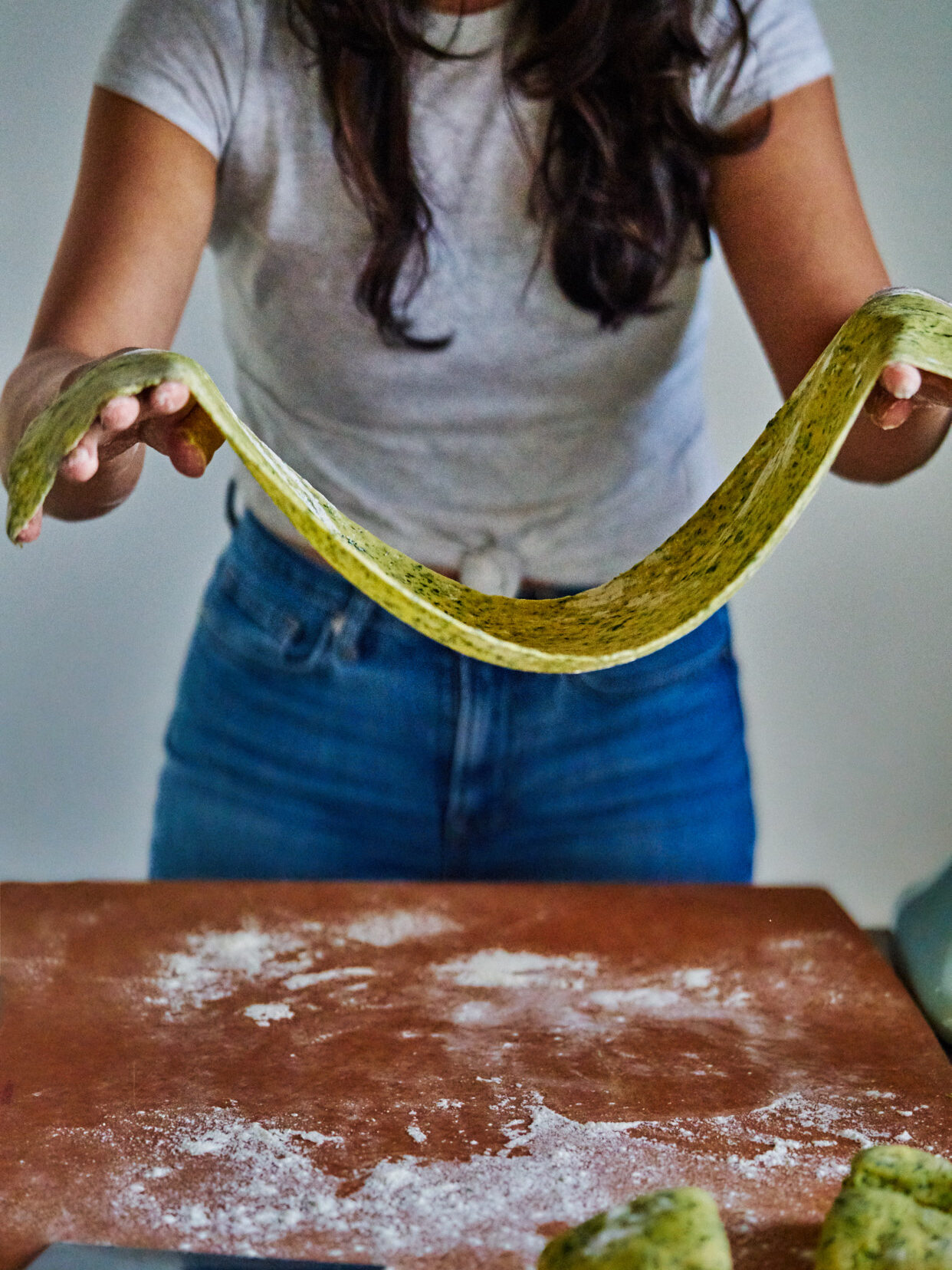 A woman stretching out a long peice of beige and green pasta above a floured counter.