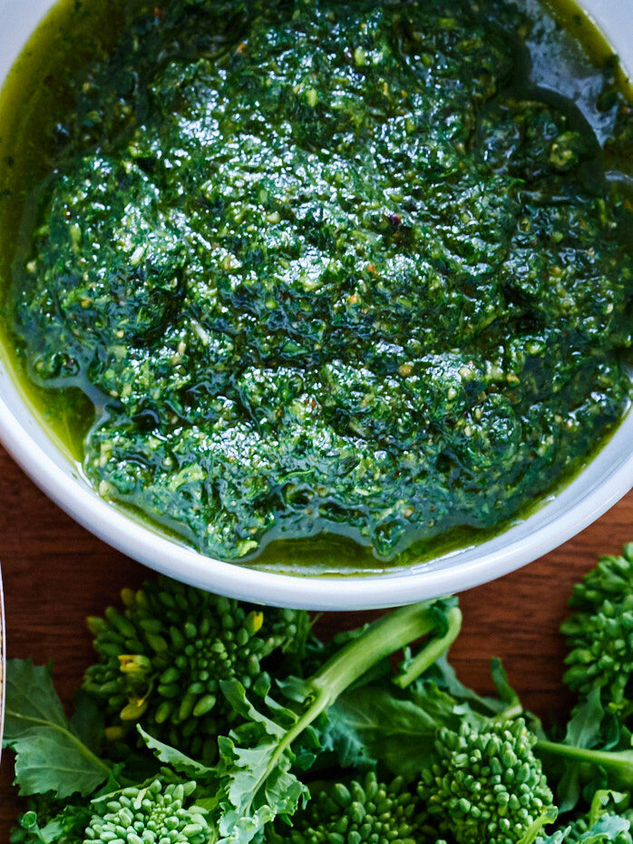 Chopped up broccoli rabe in a silver pan next to a bowl of green pesto and raw broccoli rabe.