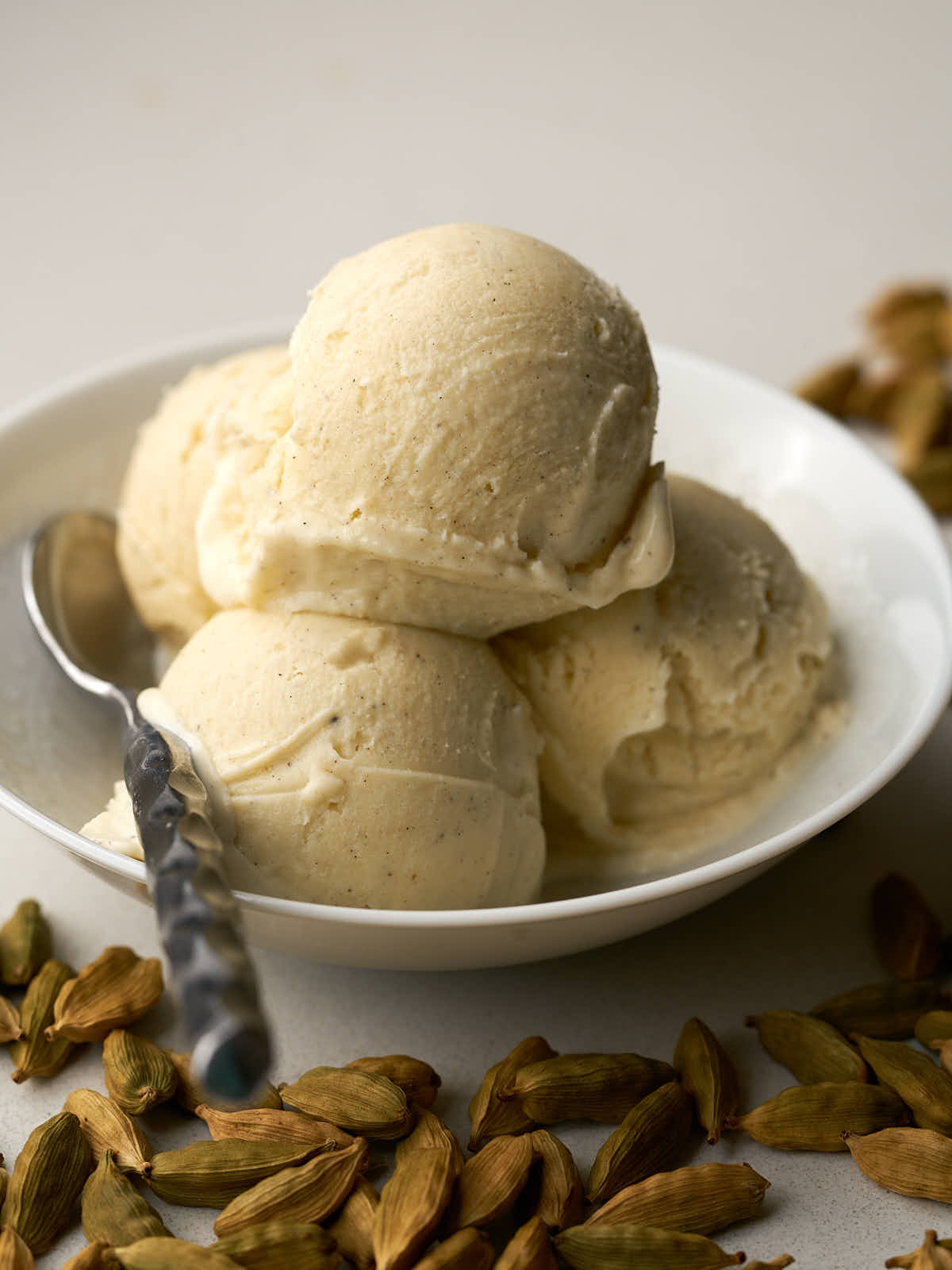 Four scoops of ice cream in a white bowl next to cardamom pods.