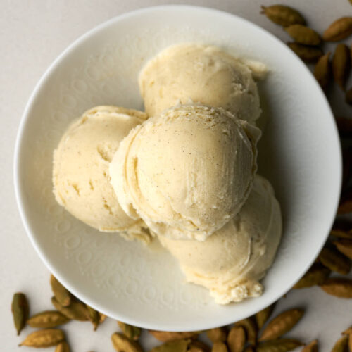 Top view of four scoops of ice cream in a white bowl next to cardamom pods.