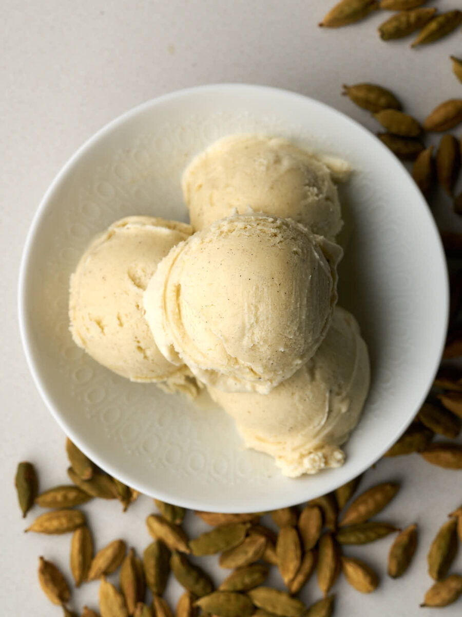 Top view of four scoops of ice cream in a white bowl next to cardamom pods.