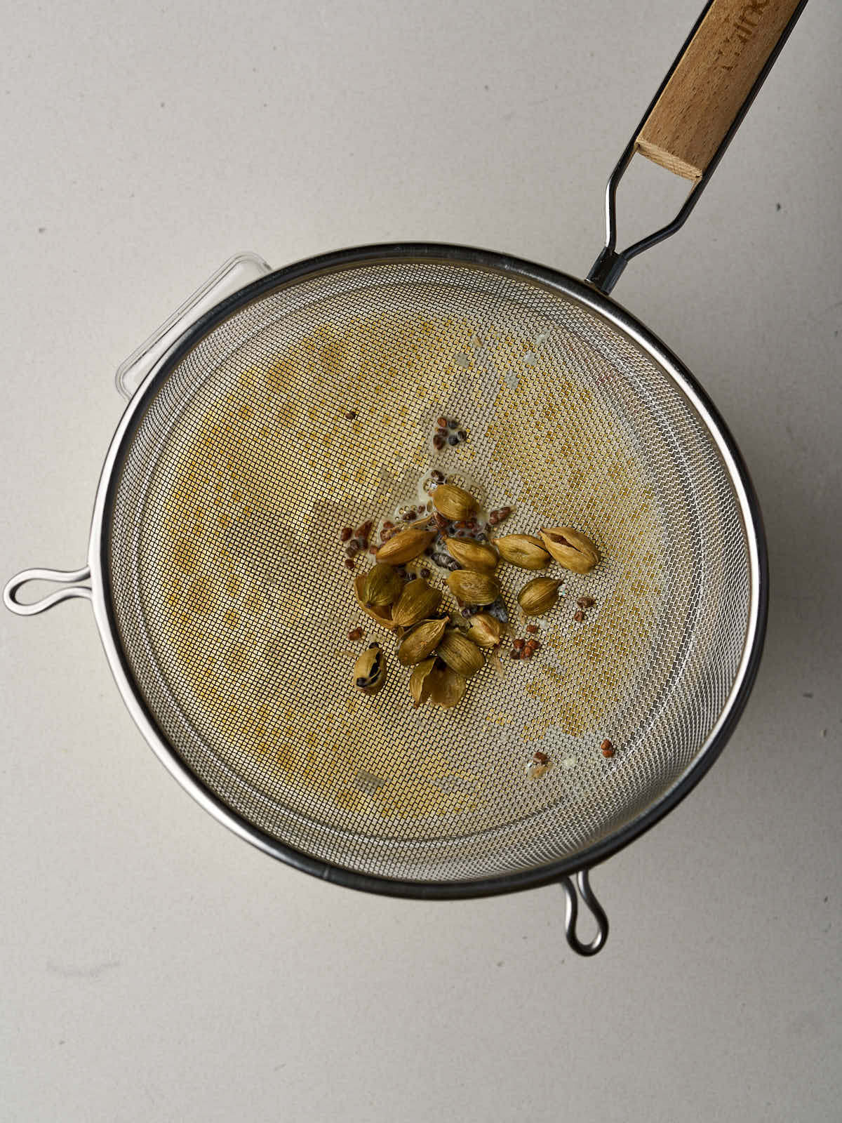 Cracked cardamom pods in a metal strainer over a plastic container with pale yellow liquid.