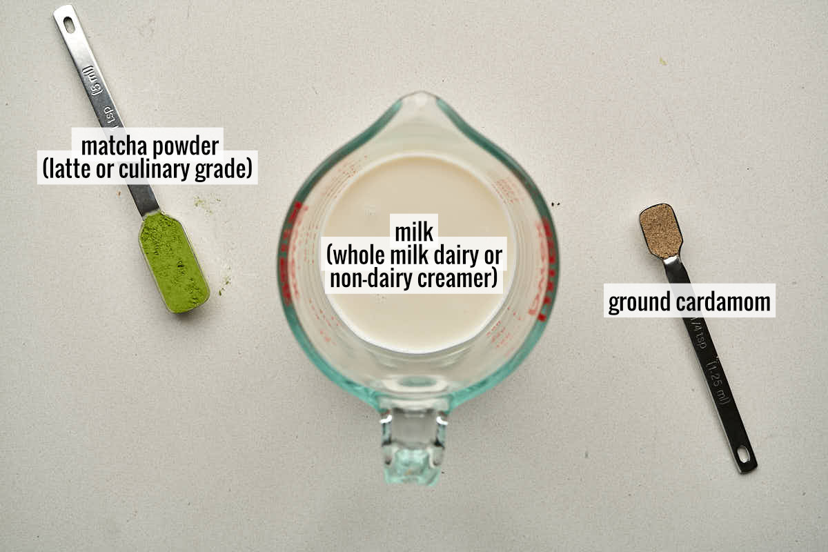 Liquid measuring cup with milk next to two measuring spoons: one with matcha powder and the other with ground cardamom.