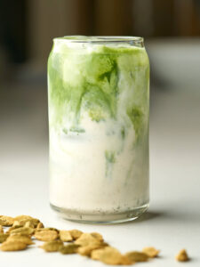 Front view of a glass soda can with an iced green match latte next to cardamom pods.