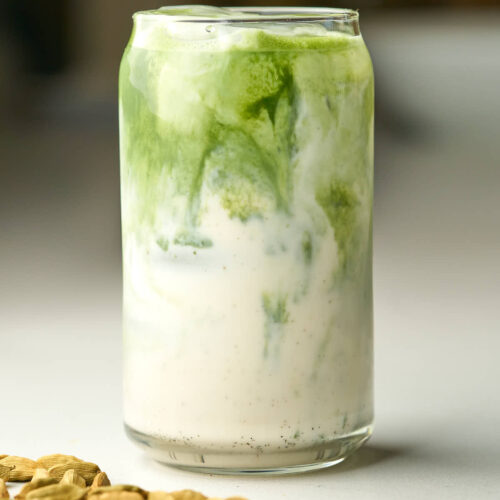 Front view of a glass soda can with an iced green match latte next to cardamom pods.