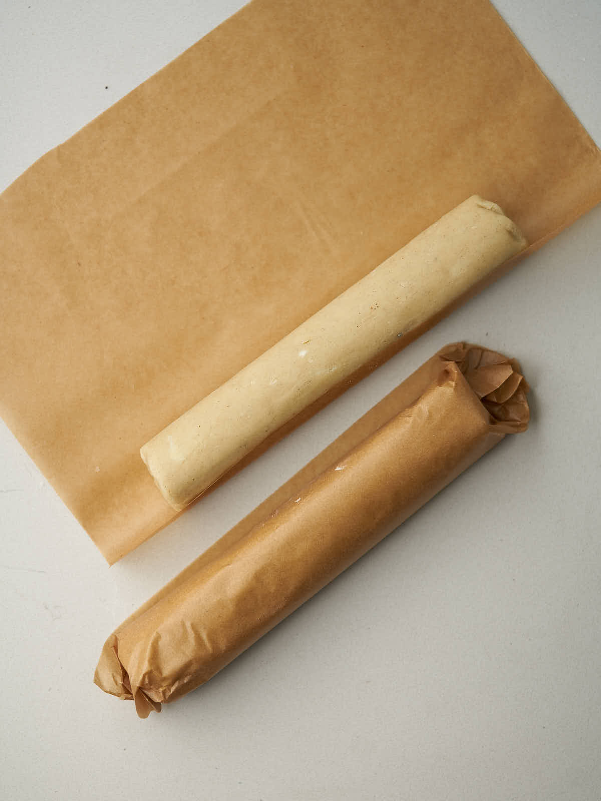 A log of shortbread dough on a piece of brown parchment paper next to a log of shortbread dough rolled in parchment paper.