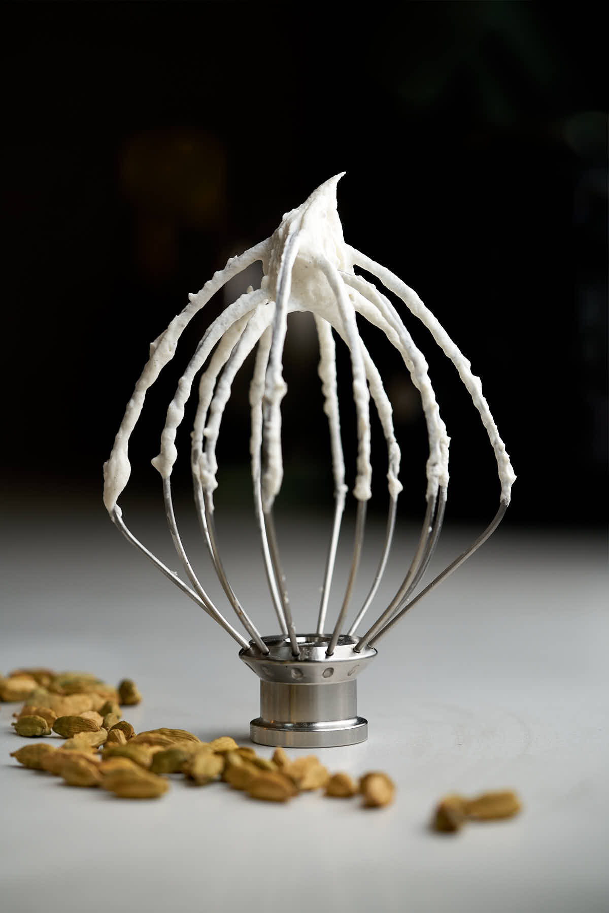 A large silver whisk attachment with whipped cream on it and cardamom pods beside it.