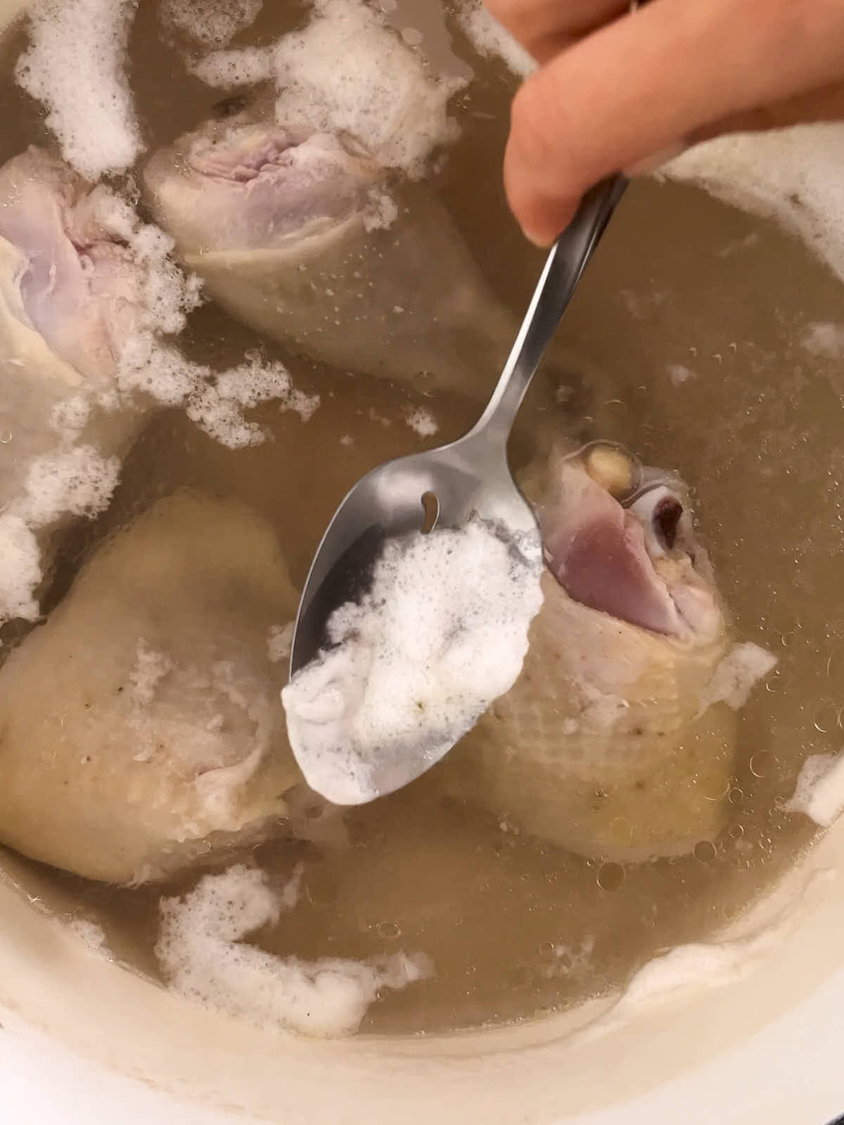 A hand using a silver spoon to remove fat from the top of chicken broth.