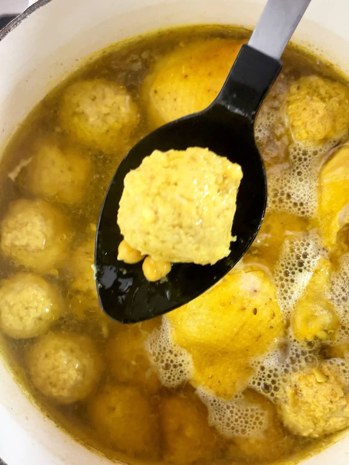 A chicken dumpling on a black cooking spoon above a pot of chicken broth.