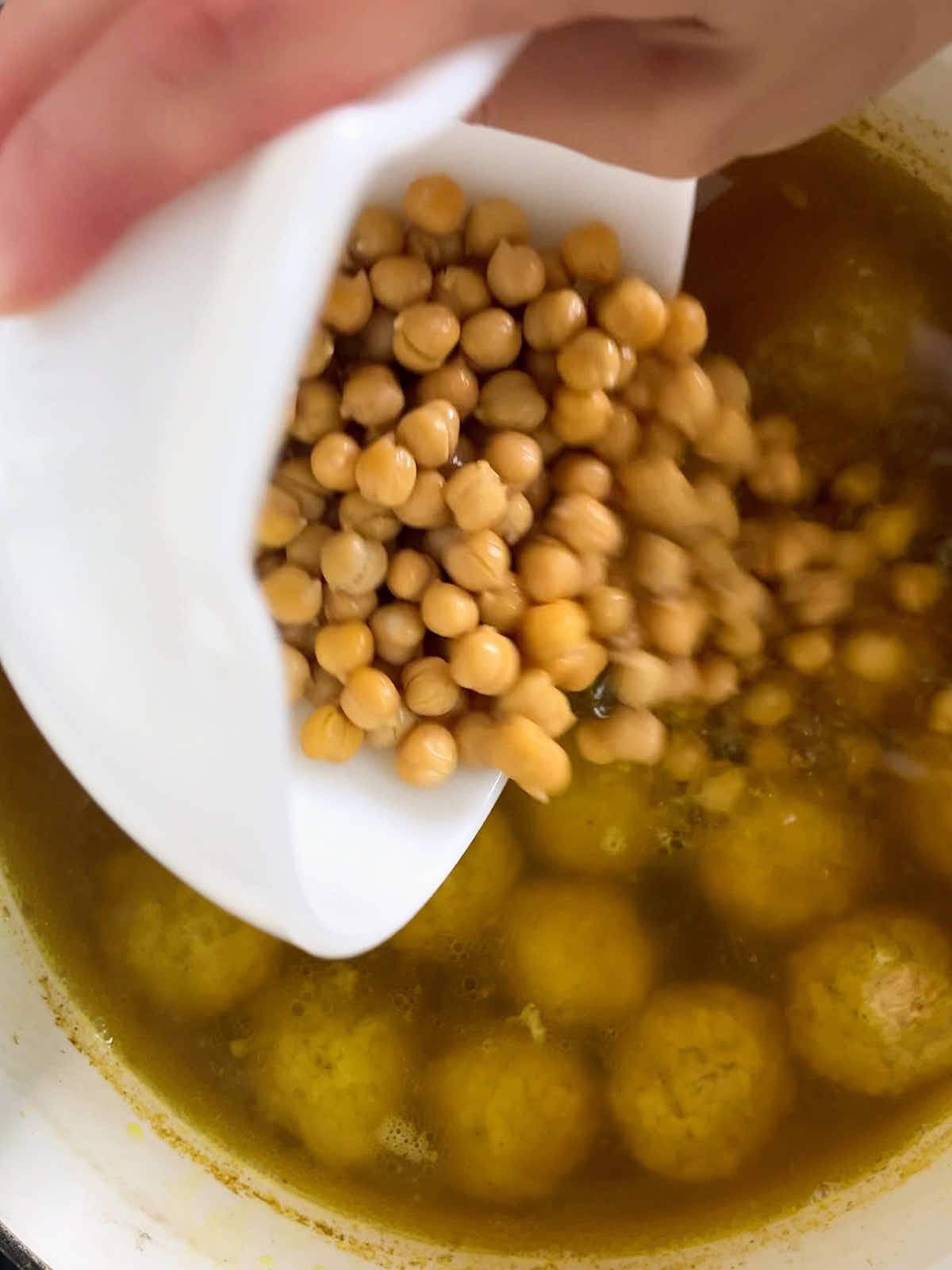 Hand pouring chickpeas into a large pot of chicken dumplings and broth.