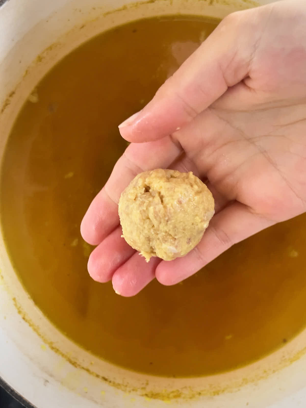 A hand holding a chicken dumpling above a stew pot.