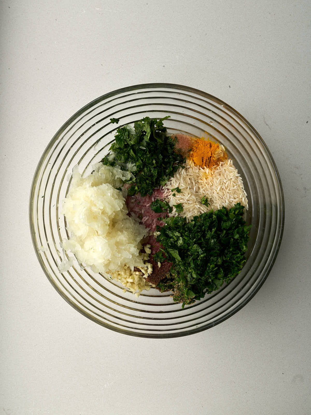 Rice, herbs, onion, and raw meat separated in a large glass bowl.