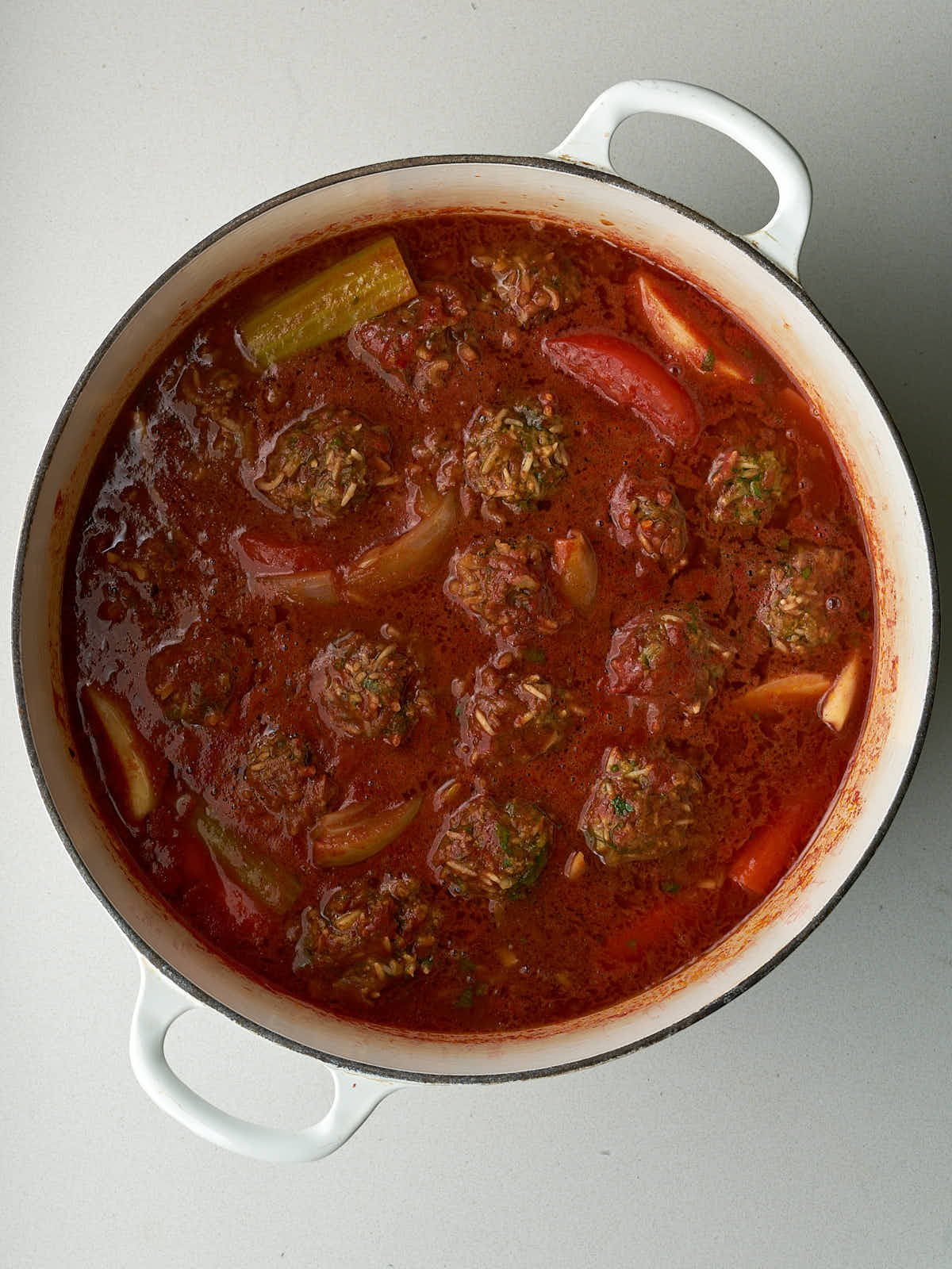 Meat and rice dumplings submerged in a large pot of red sauce with vegetables.