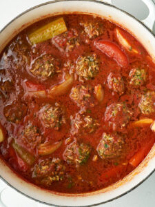 Close up of meat and rice dumplings submerged in a large pot of red sauce with vegetables.