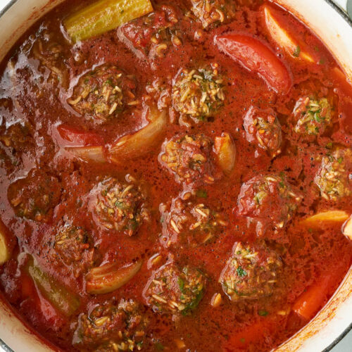 Close up of meat and rice dumplings submerged in a large pot of red sauce with vegetables.