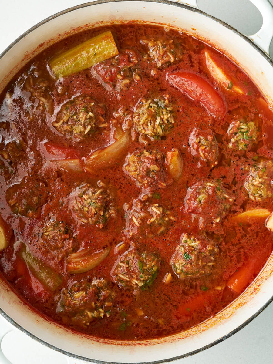 Close up of meat and rice dumplings submerged in a large pot of red sauce with vegetables.