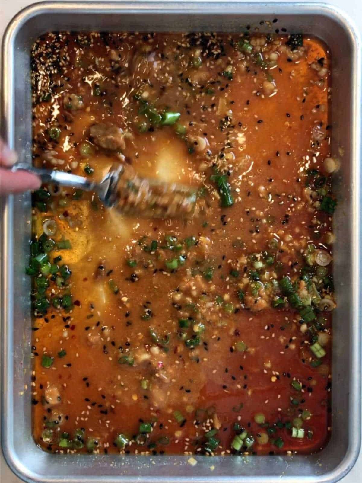 A hand using a fork to stir a marinade on a baking sheet.