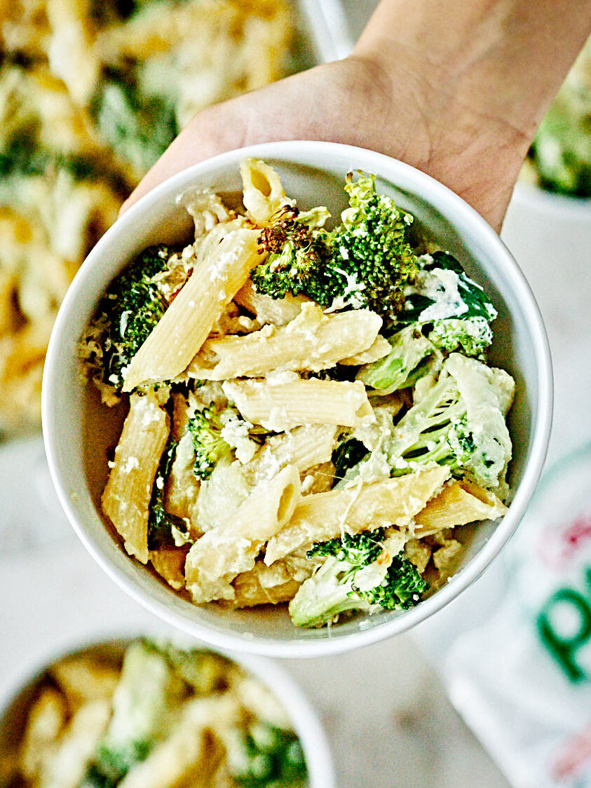 A hand holding a white bowl of penne pasta with broccoli and white sauce.