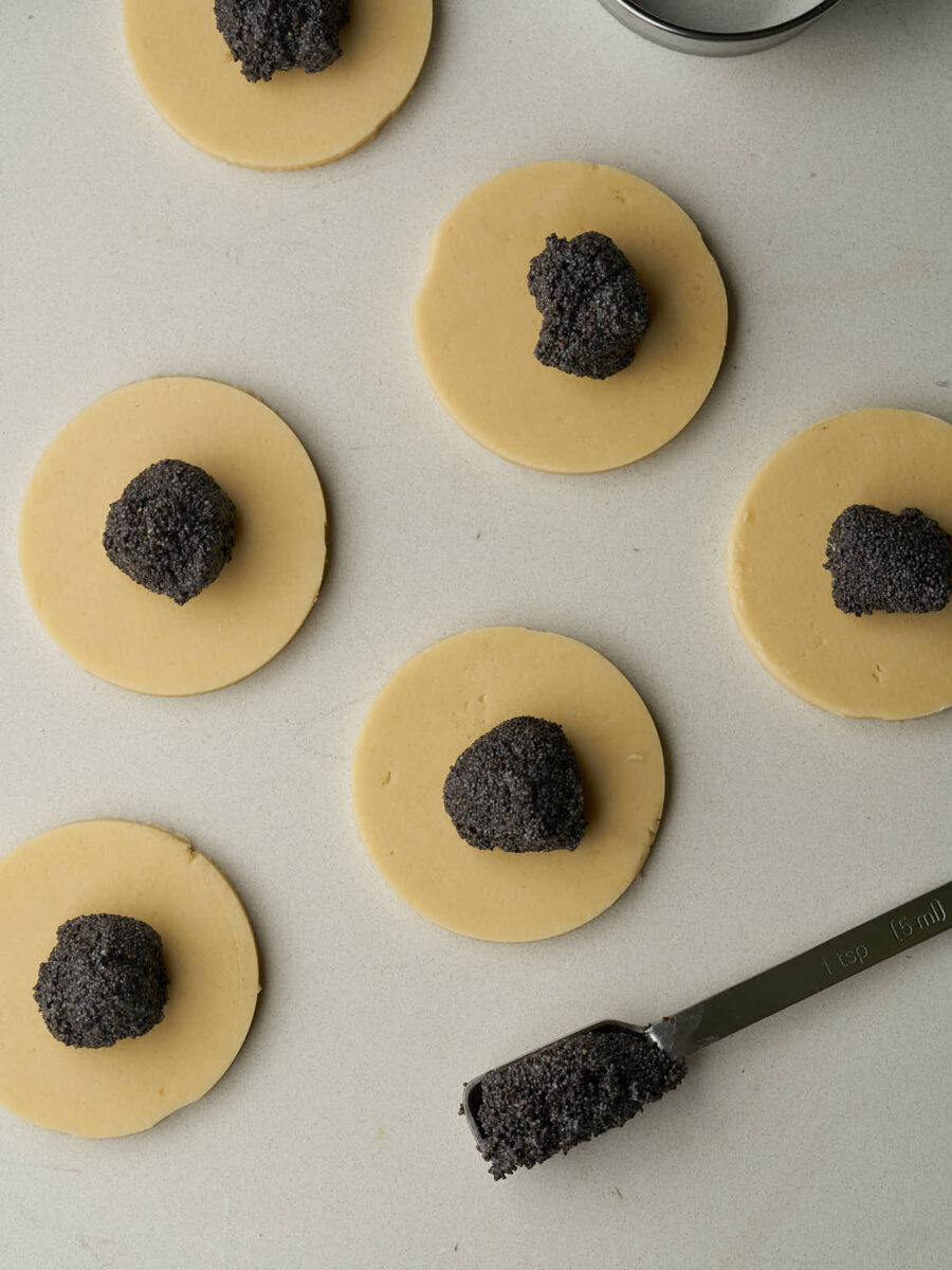 Circles of dough with dollops of dark grey poppyseed filling in the centers next to a measuring teaspoon filled with poppyseed filling.