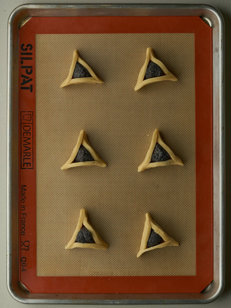 Triangular cookies filled with poppyseeds on a silicone baking mat with a red rim.