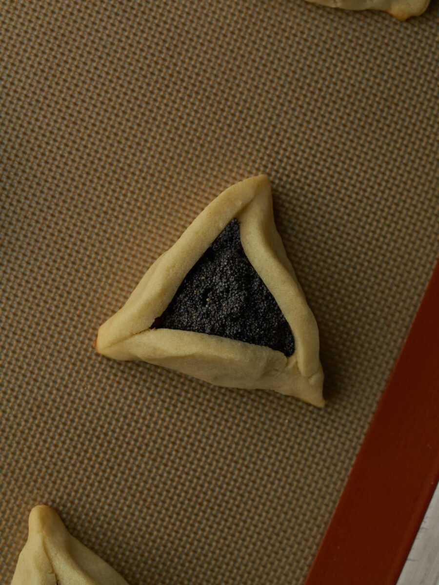 One triangular cookie with black poppyseed filling on a silicone baking mat lined baking sheet.