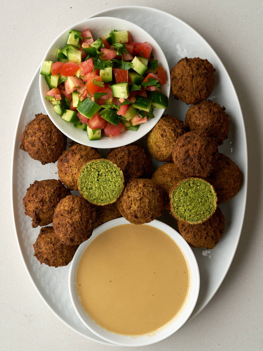 Falafel on an oval platter with a small bowl of tahini and another small bowl of cucumber tomato salad and two falafel halves with green insides.