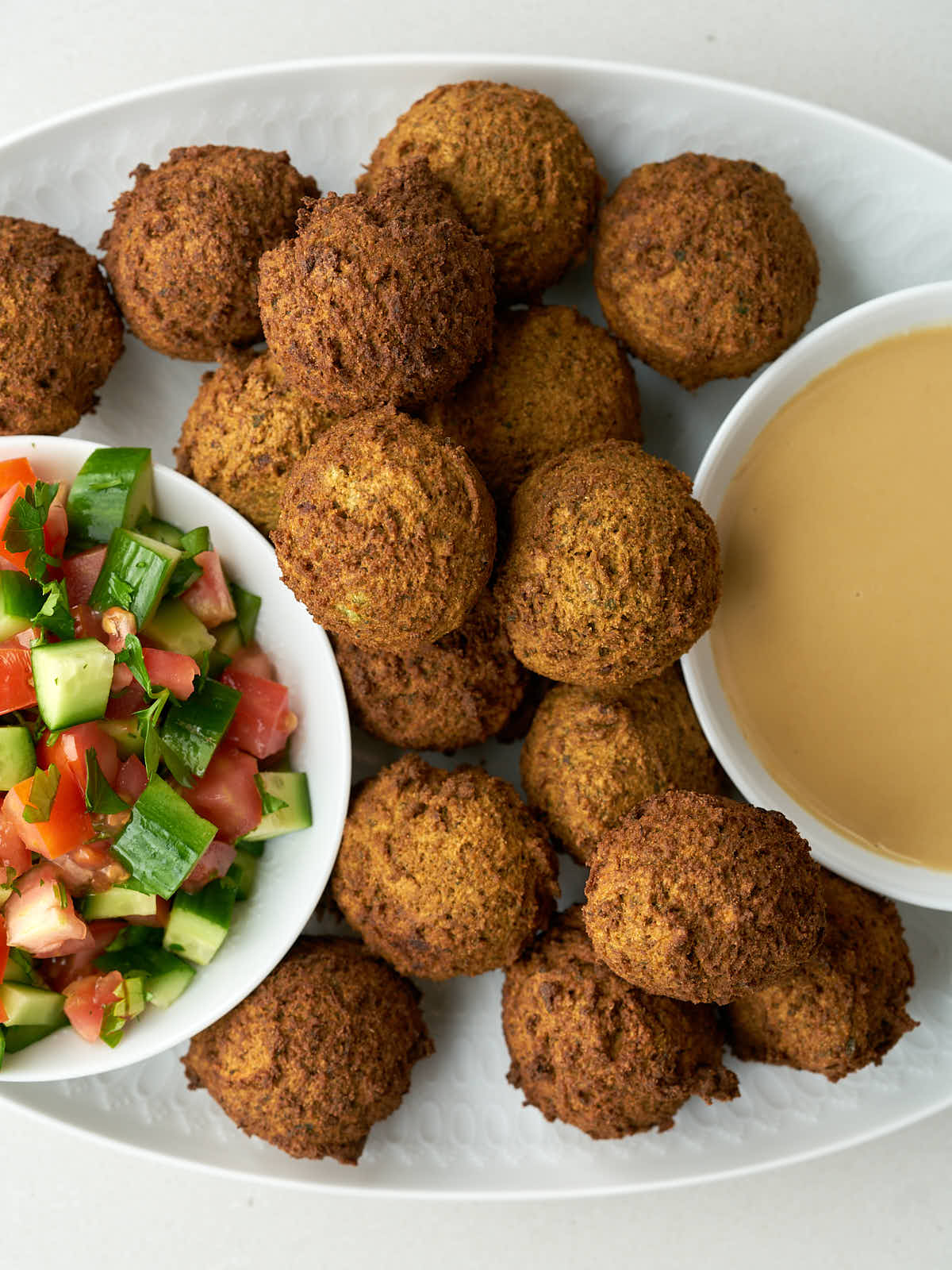 Falafel on an oval platter with a small bowl of tahini and another small bowl of cucumber tomato salad.